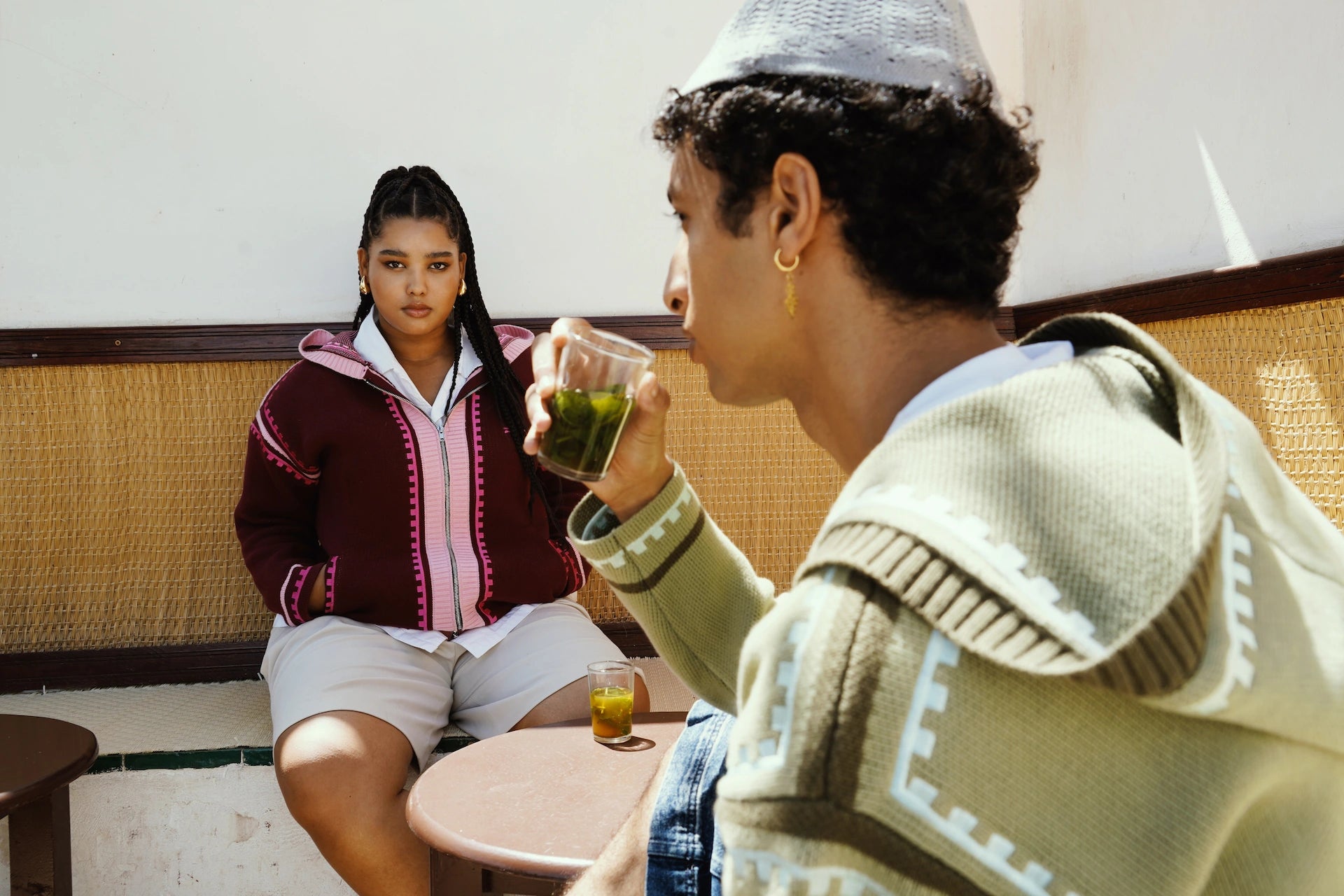 A girl and boy wearing R'MILI Berbere Knitwear Jackets in burgundy and green, drinking tea in Rabat